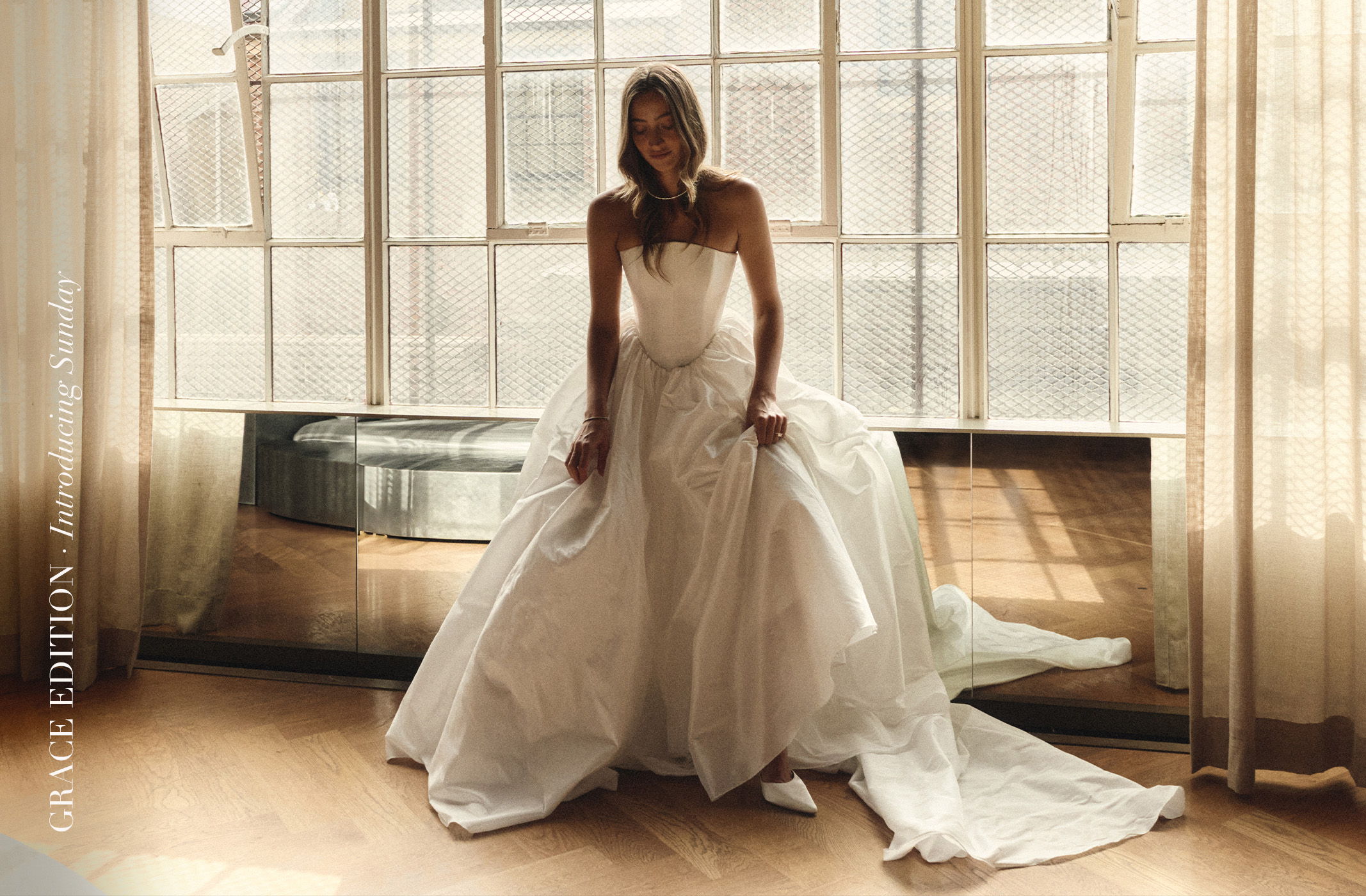 Woman in a wedding dress sitting by large windows in soft light.