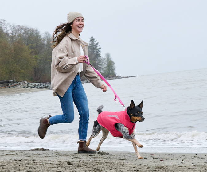 Woman joyfully runs alongside her loyal dog with a coat and a leash on a beach