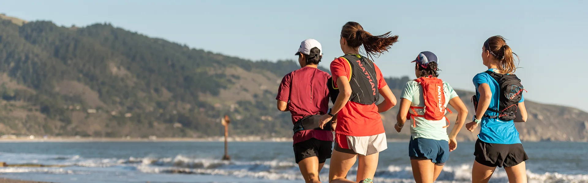 Group of runners wearing Nathan hydration packs and vests while running down the beach with waves rolling in