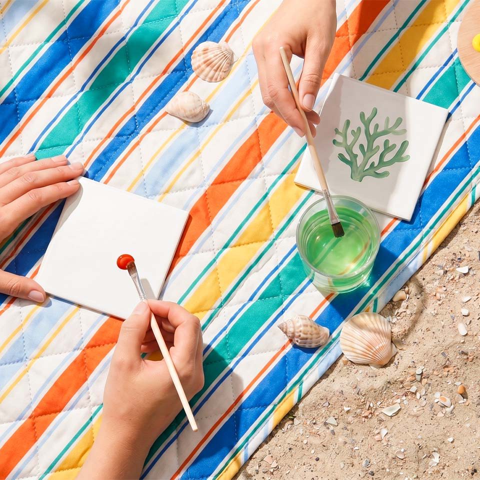 Hands painting on paper with shells nearby on a colorful striped blanket.