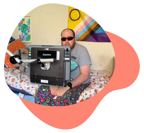 A person sitting on a bed using a Grid Pad 13 communication device. with a pride flag in the background.