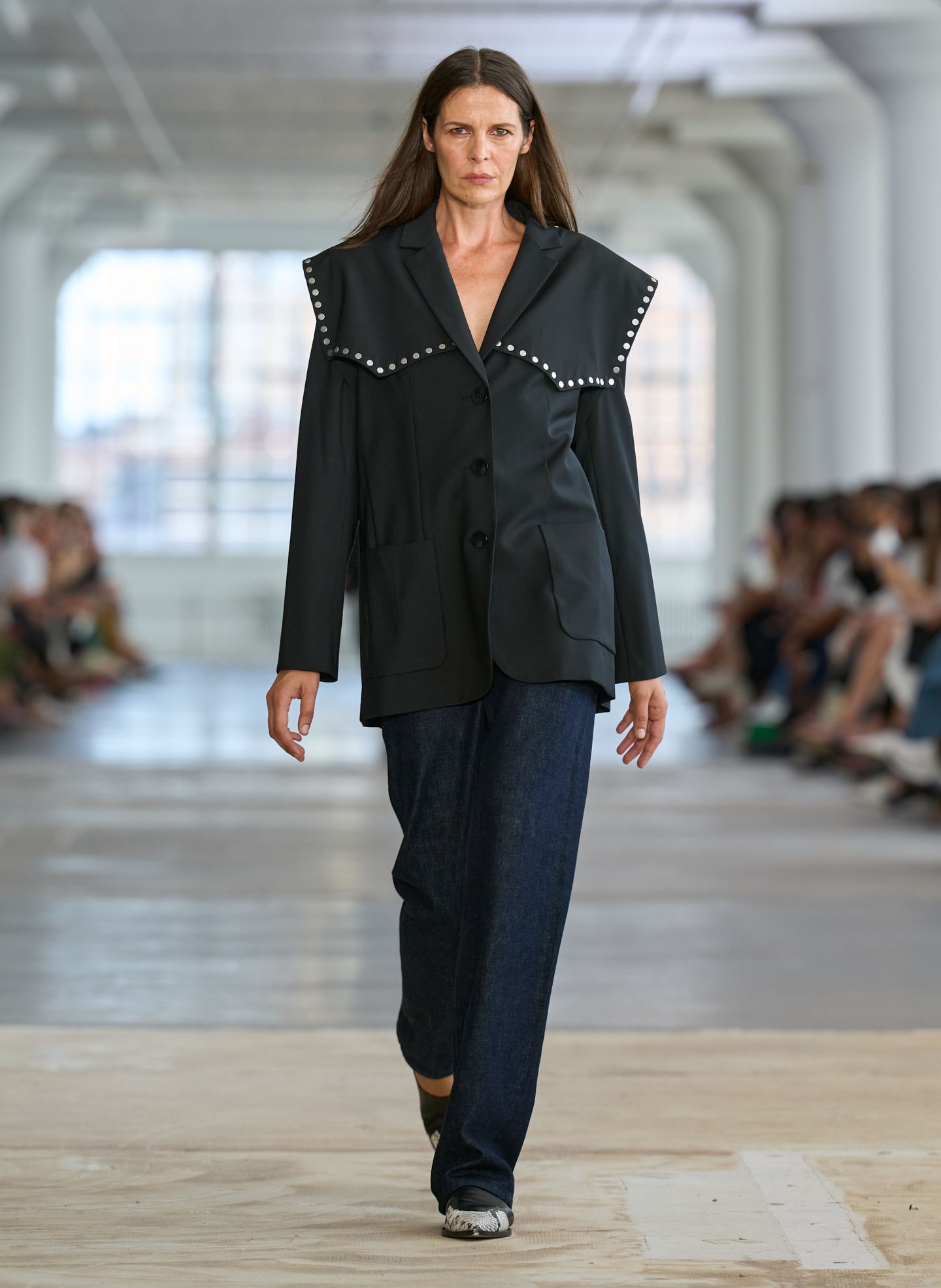 Model walking down runway wearing oversized jacket and dark pants.
