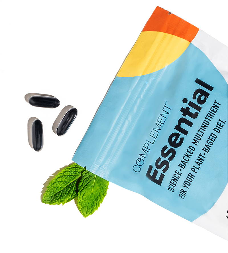 Complement Essential vegan multivitamin pouch next to mint leaves and black capsules. 