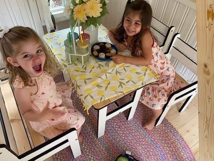  2 girls have a lemonade party inside a black-and-white wooden playhouse with kids furniture by WholeWoodPlayhouses