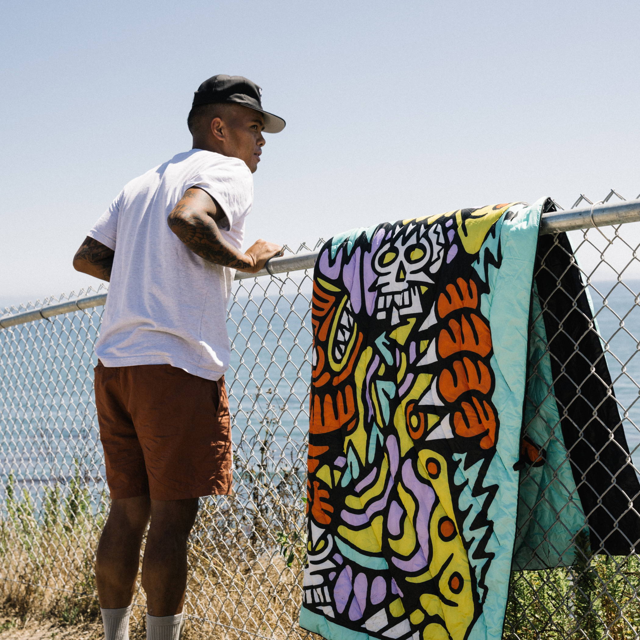 DJ Javier enjoys the ocean view, leaning over a wire fence with the limited edition In the Balance Original Puffy Blanketb draping over it.