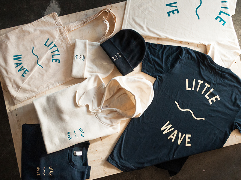 Flat lay of cream and navy Little Wave sweat shirts, tees and beanies printed and embroidered with the Little Wave logo and wave motif