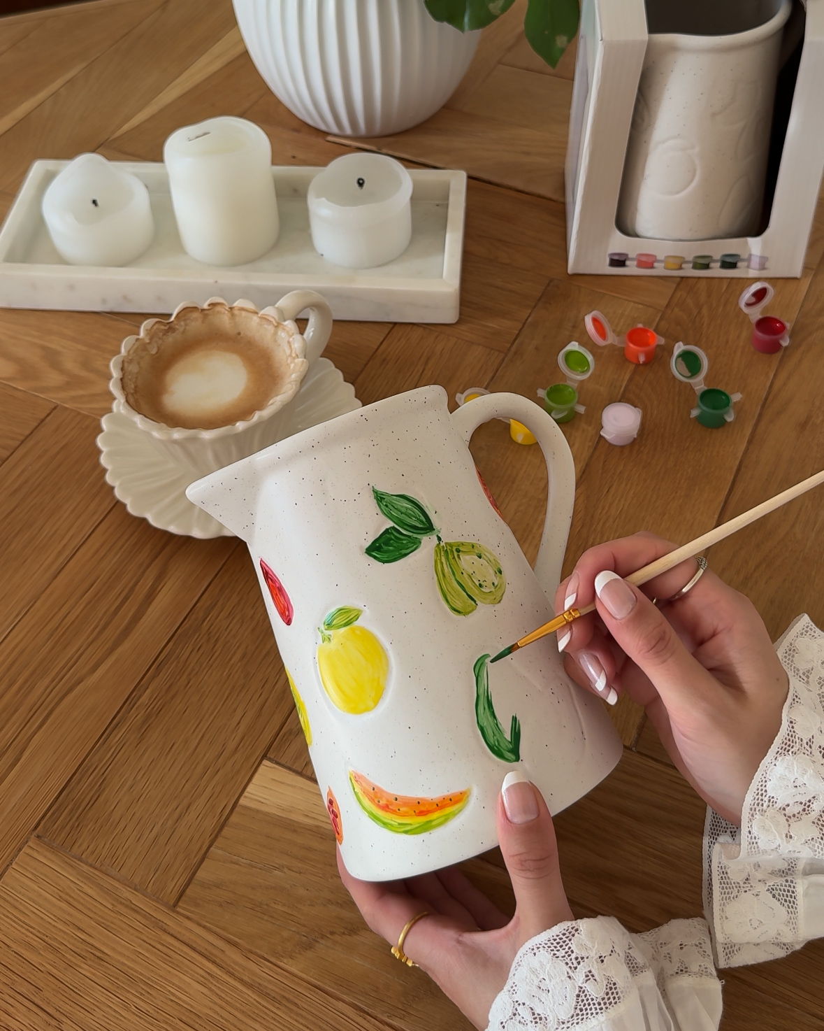 Person painting a colorful pitcher on a wooden table with coffee and candles.