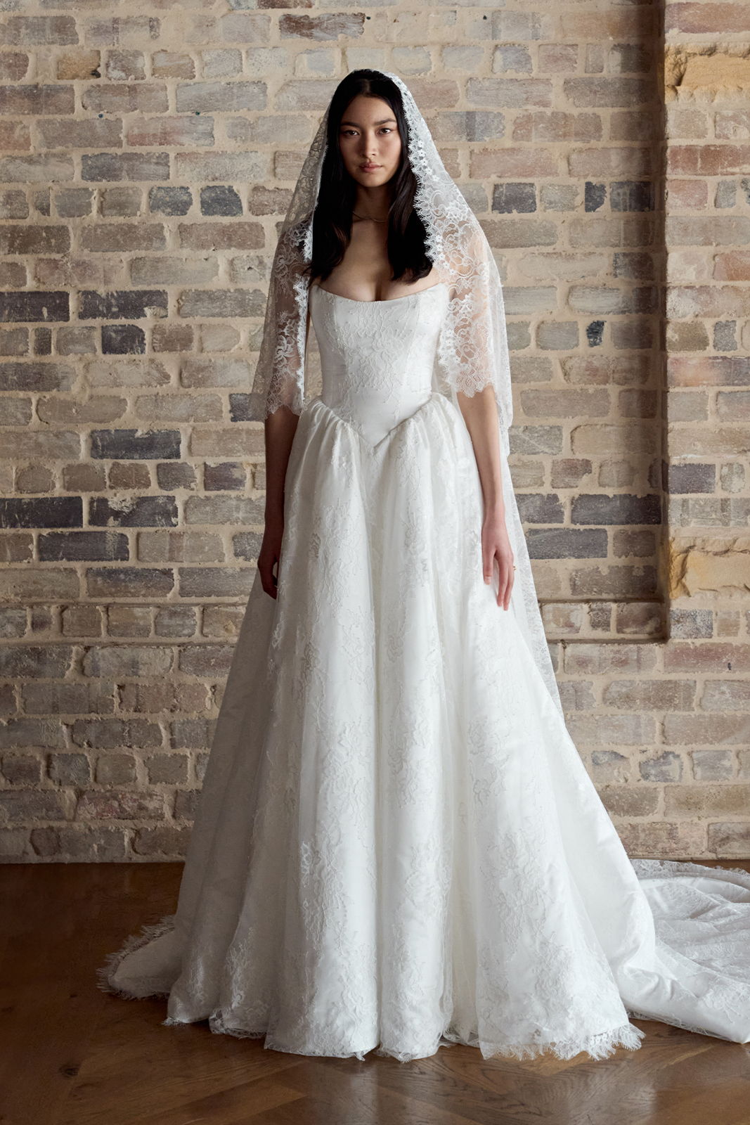 A woman in a white wedding dress with a veil standing against a brick wall.
