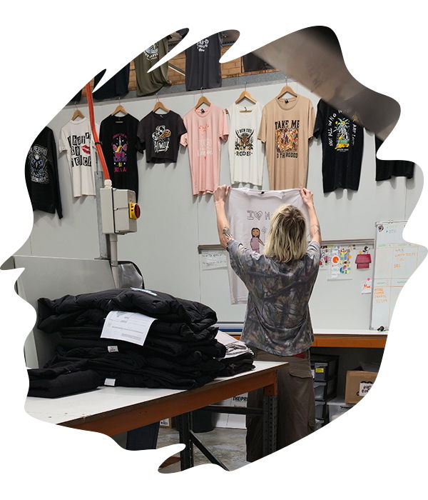 Staff member checking finished printed t-shirtswith various shirts hanging on a wall rack in the production studio, with folded black garments stacked on a table below