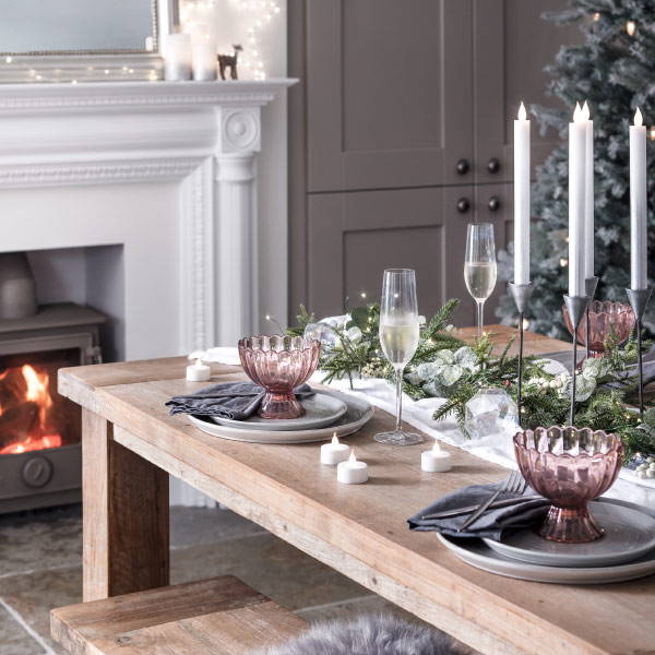 A wooden dining table with LED taper candles and a garland table runner with a fireplace and Christmas tree in the background.