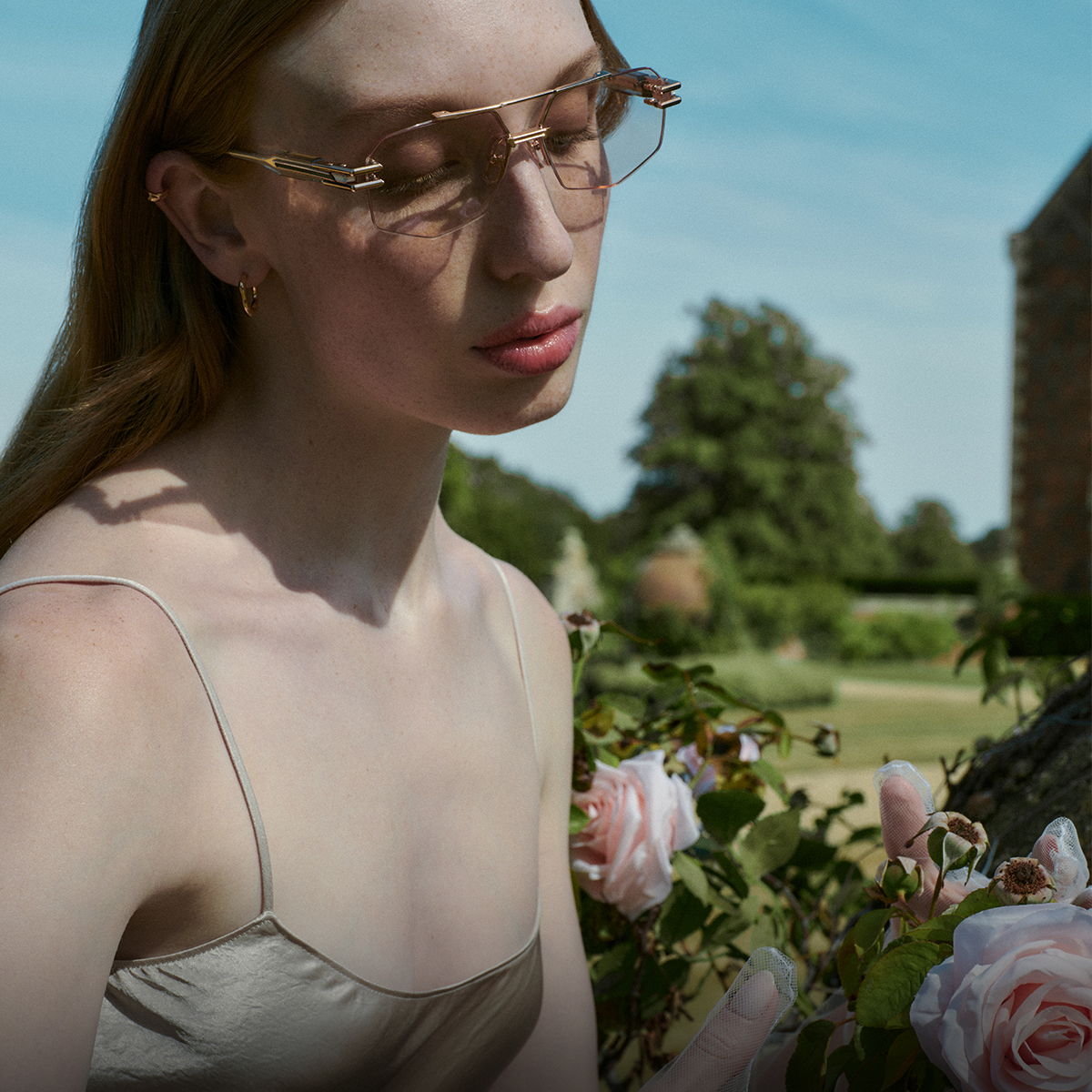 A person with glasses sitting near pink roses in a garden.