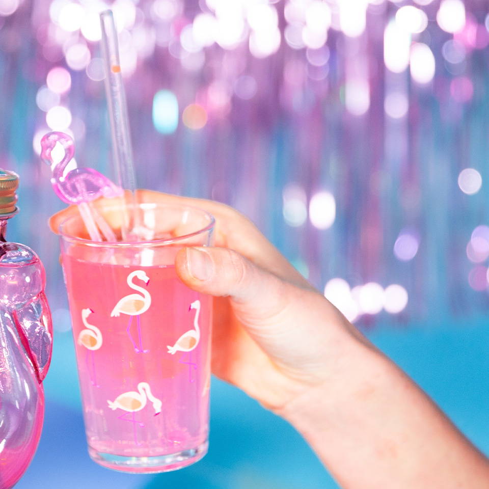 Close-up of a hand holding a pink glass with a flamingo straw, set against a festive, glittery background
