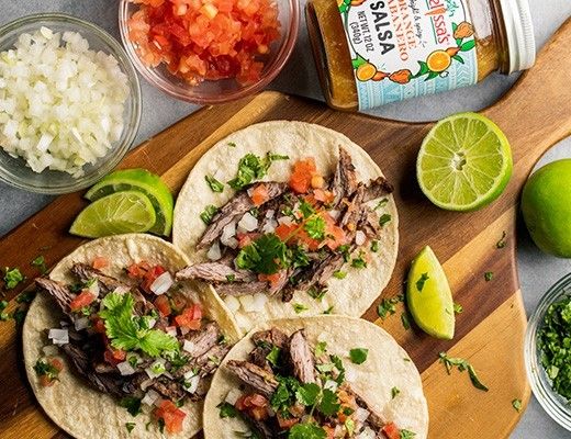 Tacos with meat, salsa, lime, onions, and cilantro on a wooden board.