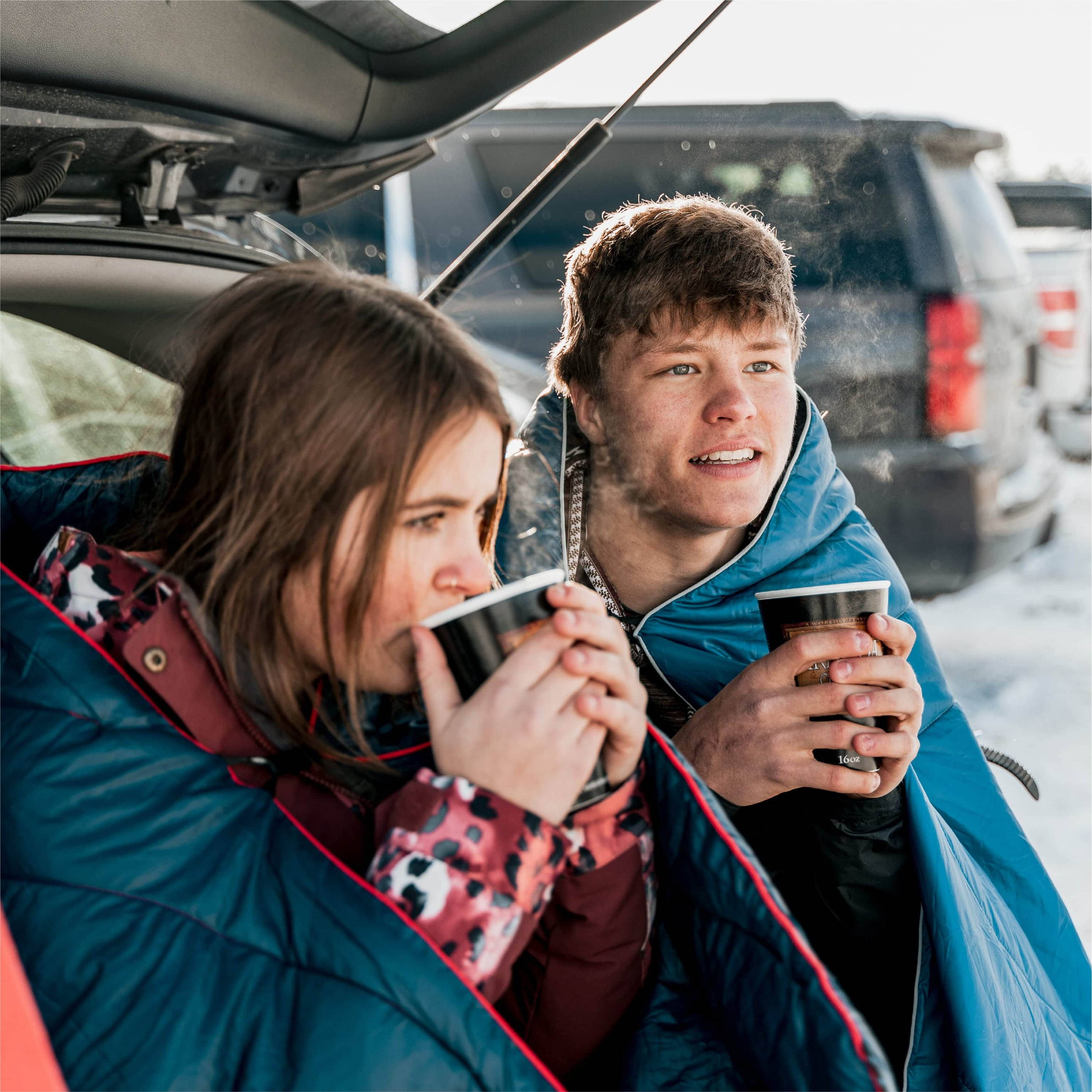 Man And Woman Drinking Coffee Outside Of Back Of Car While Wrapped In Original Puffy Blankets