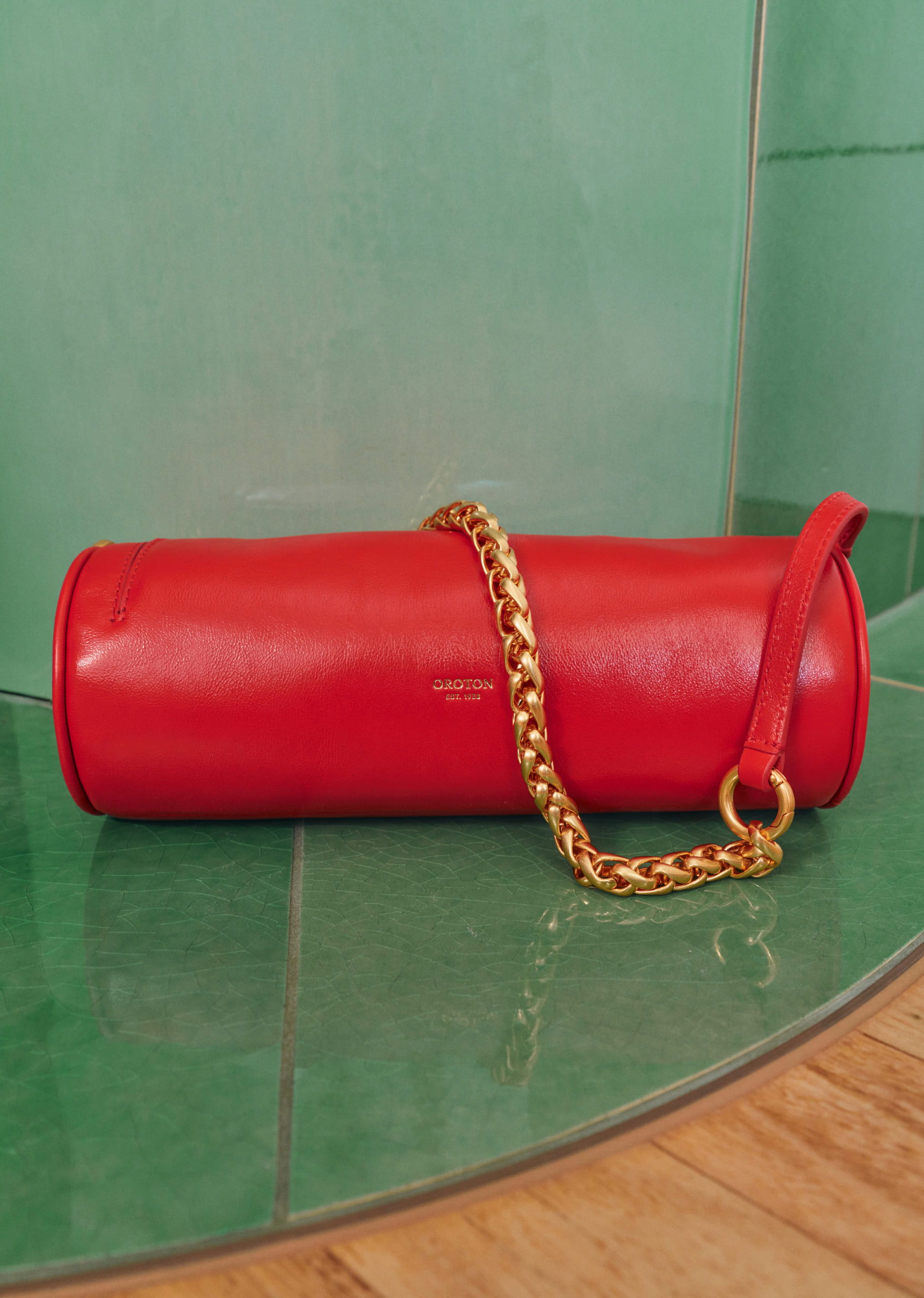 Red cylindrical handbag with a gold chain strap on a green surface.