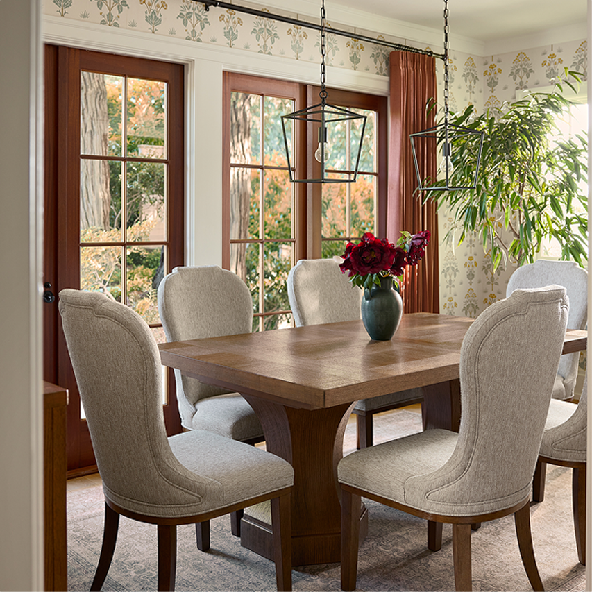 Wood dining table with upholstered chairs positioned near large windows and greenery.