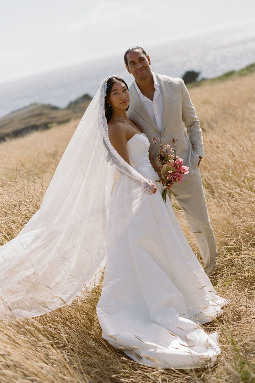 Couple in wedding attire standing in a field with ocean in the background.