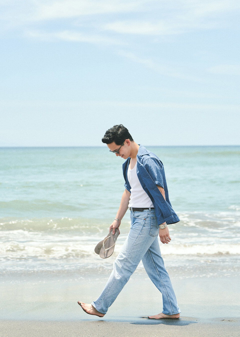 A person walking along the beach, holding a hat, near the water.