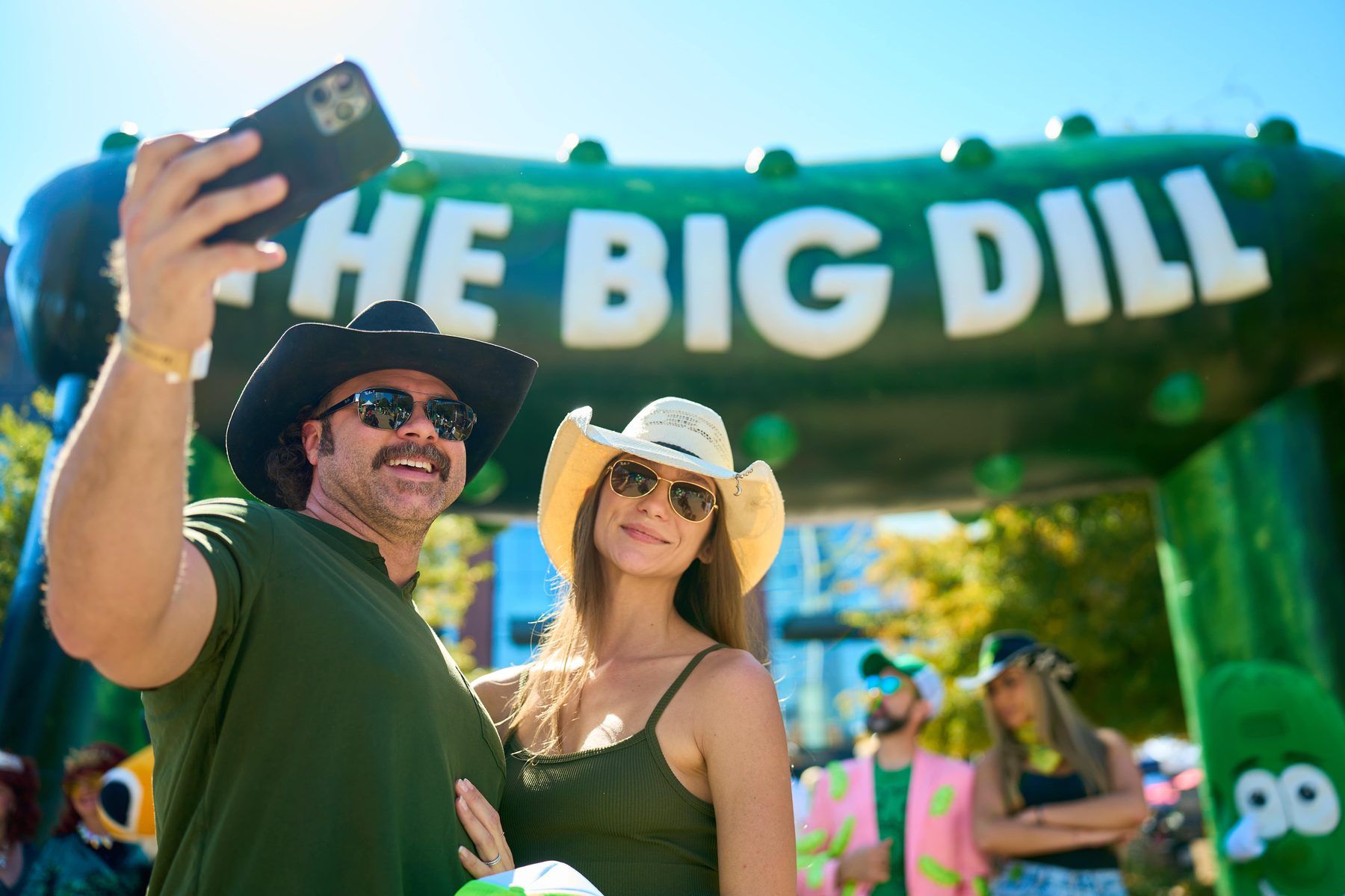 Two attendees in cowboy hats taking a selfie under The Big Dill 30-foot pickle arch at Globe Life Field in Arlington Texas 2025.