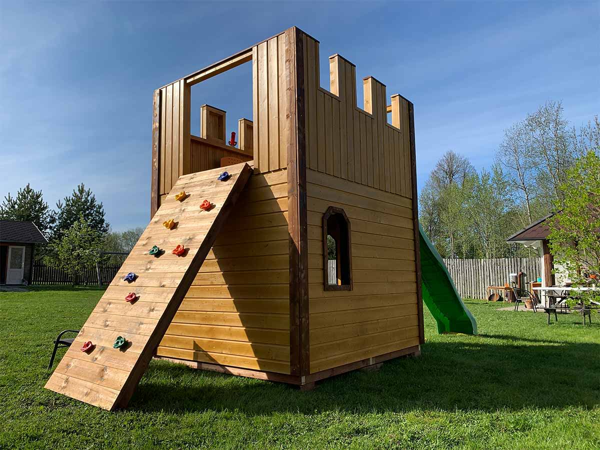 Custom Outdoor Playhouse Wooden Castle with slide and climbing wall on grass in front of green trees by WholeWoodPlayhouses