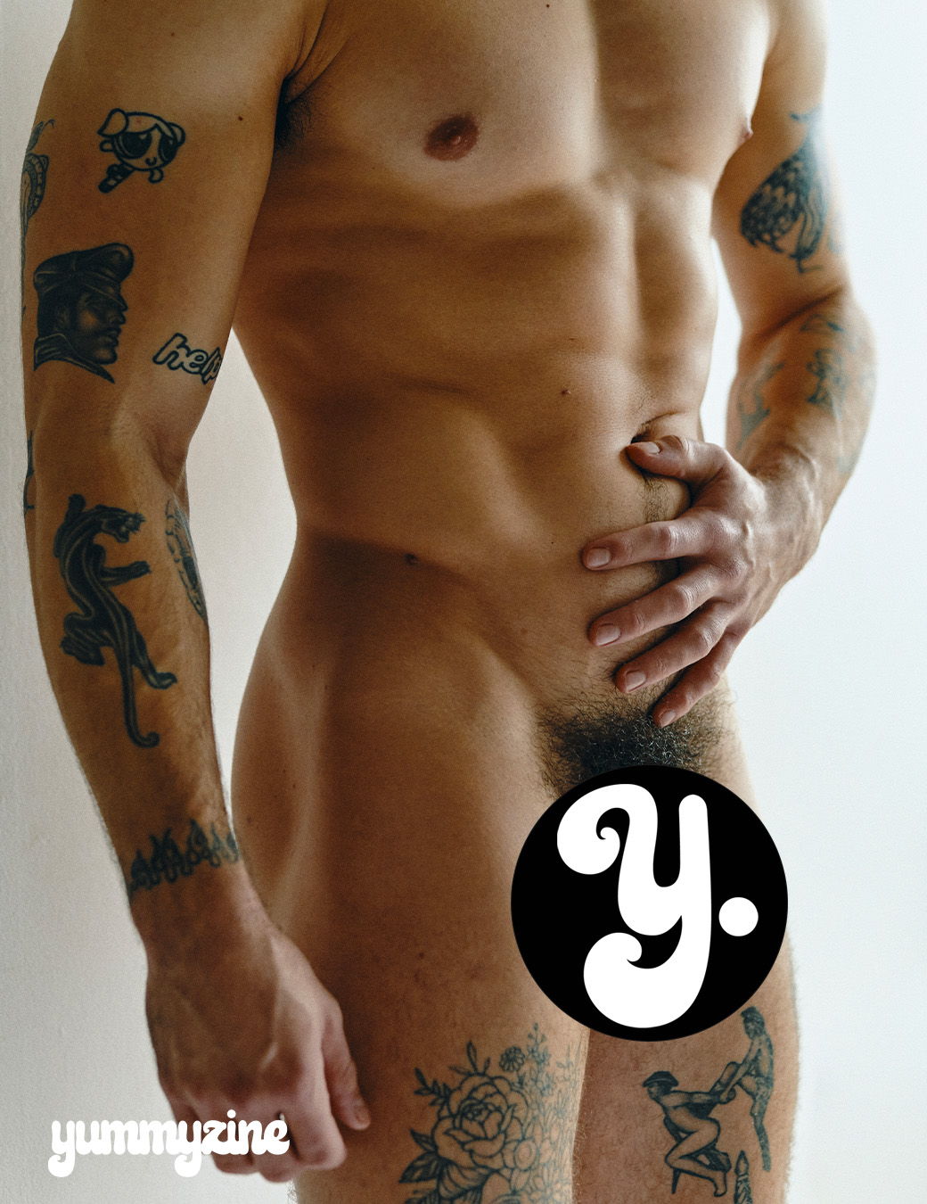 Nude male model with tattoos, covering private area with hand.