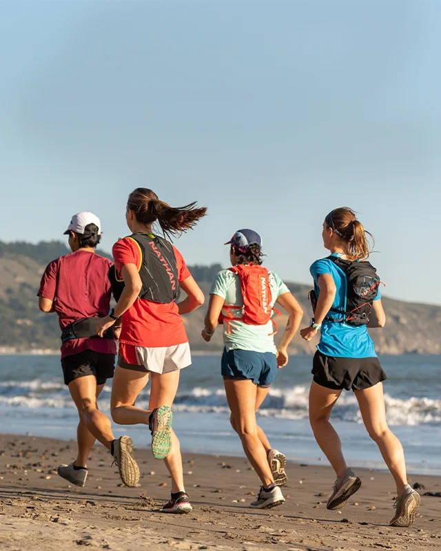 Group of runners wearing Nathan hydration packs and vests while running down the beach with waves rolling in