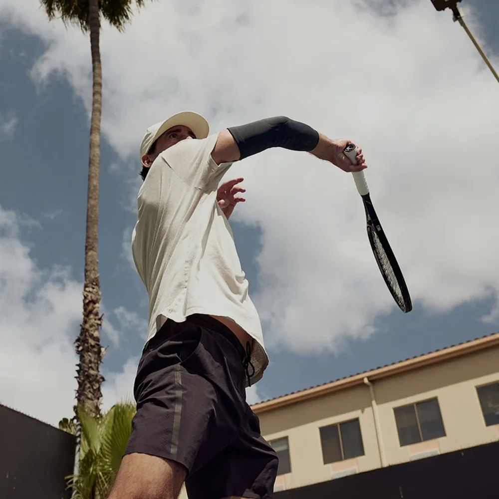 Tennis player preparing to serve with palm trees and clouds in the background.