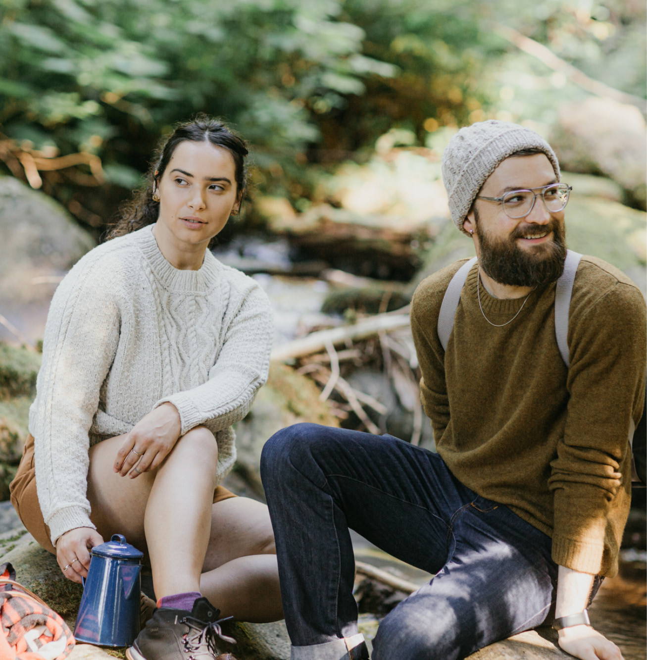 A male and a female model sitting next to each other on large river rocks in the forest.