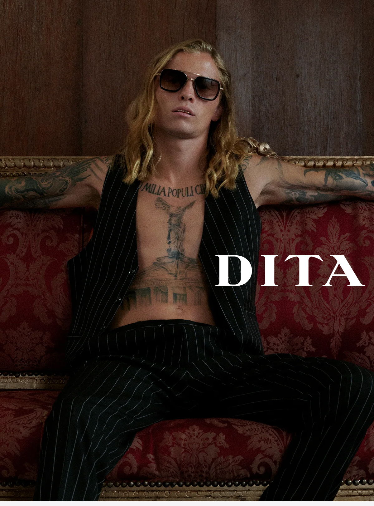 A tattooed man in sunglasses lounging on a red velvet couch.