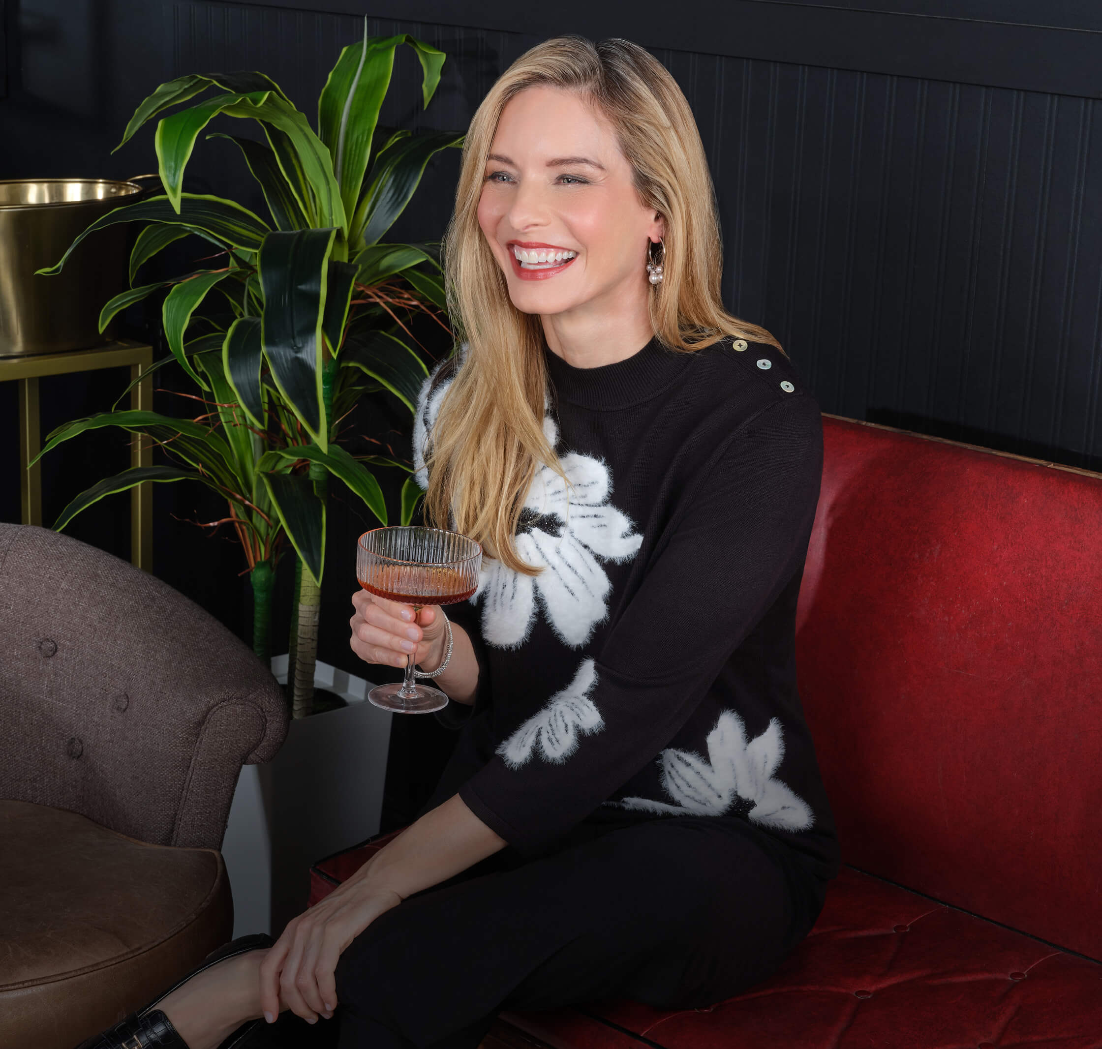 Smiling woman in a floral sweater holding a drink, seated on a couch.