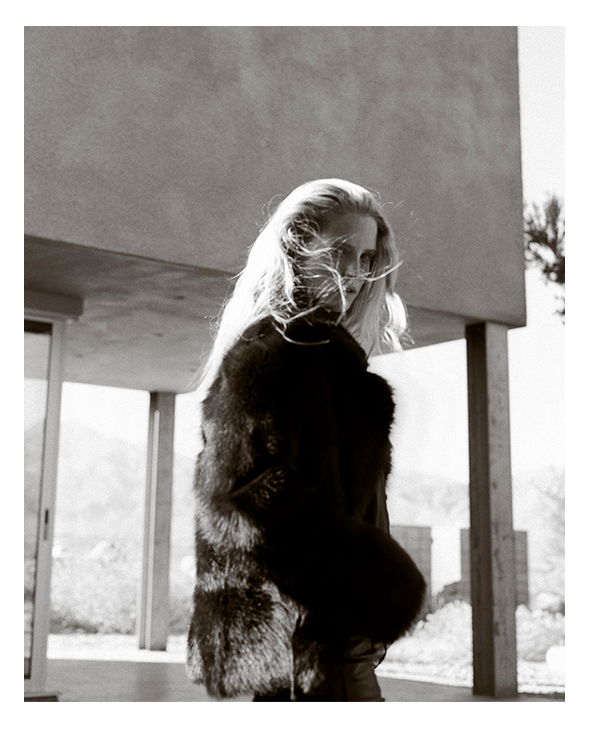 Woman with long hair in a fur coat, standing outdoors in black and white.