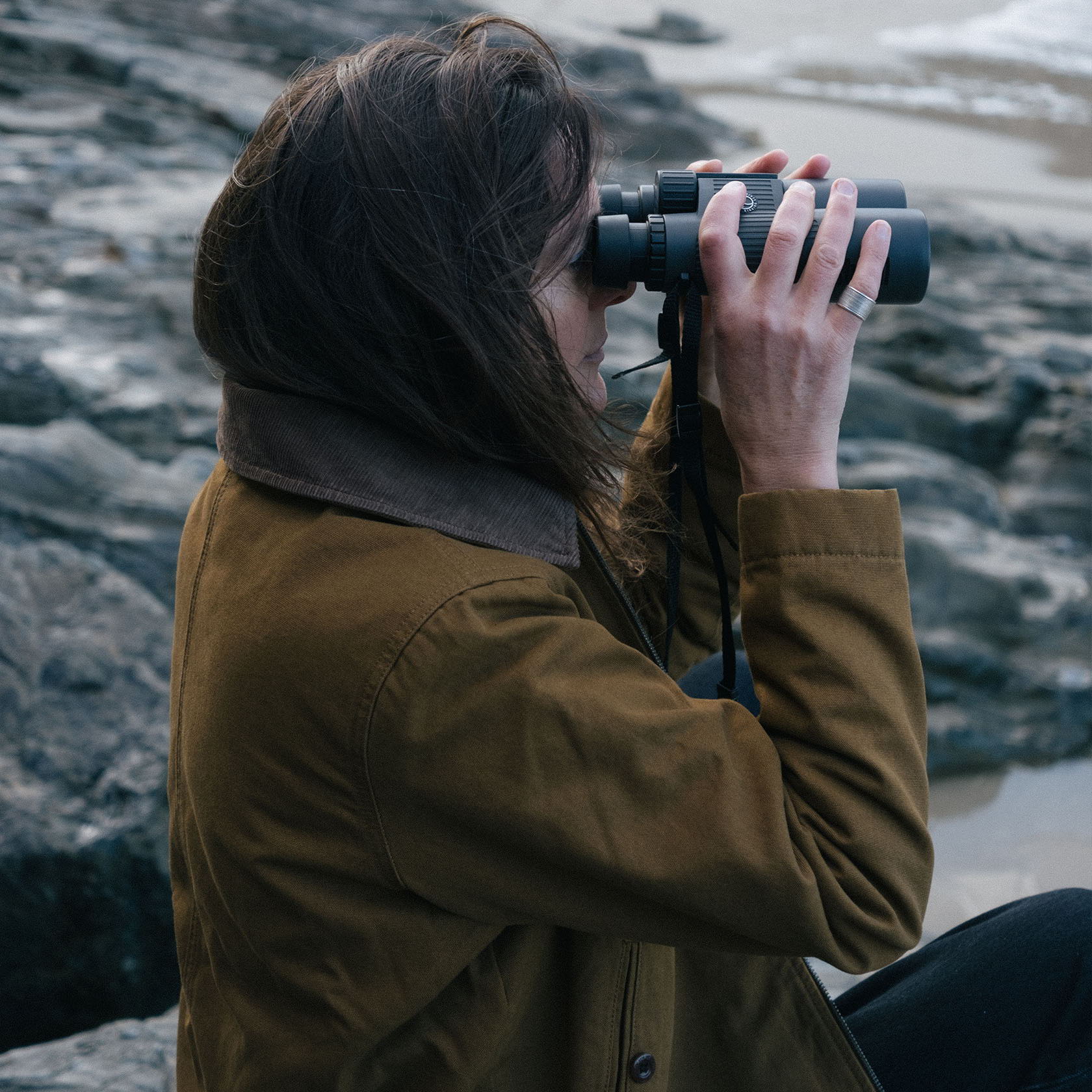 Person observing with binoculars while sitting on rocks by the shore.