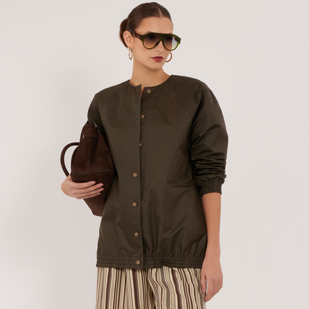 Woman wearing sunglasses and a brown jacket, holding a bag, posing.