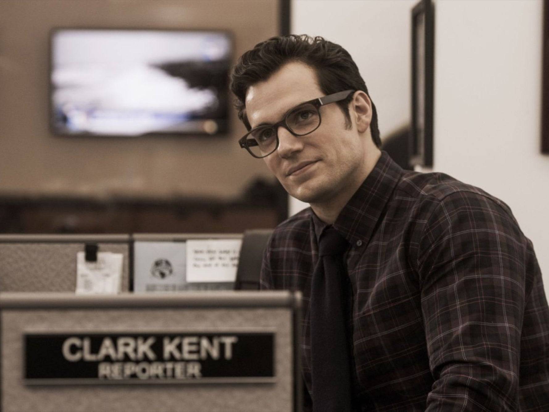 Actor Henry Cavill posing as clark kent the reporter wearing a patterned shirt, tie and standing in his office as a reporter