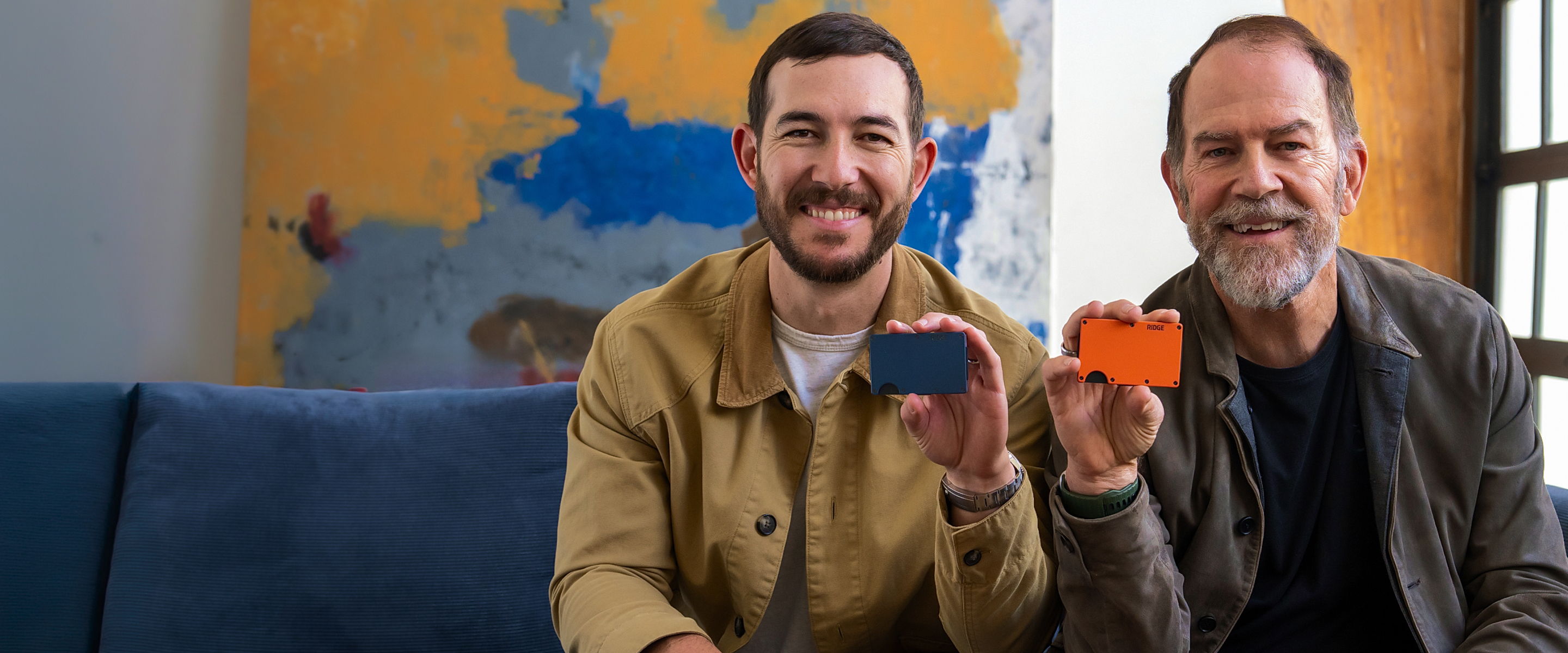 Two men smiling and holding colored objects, sitting on a couch.