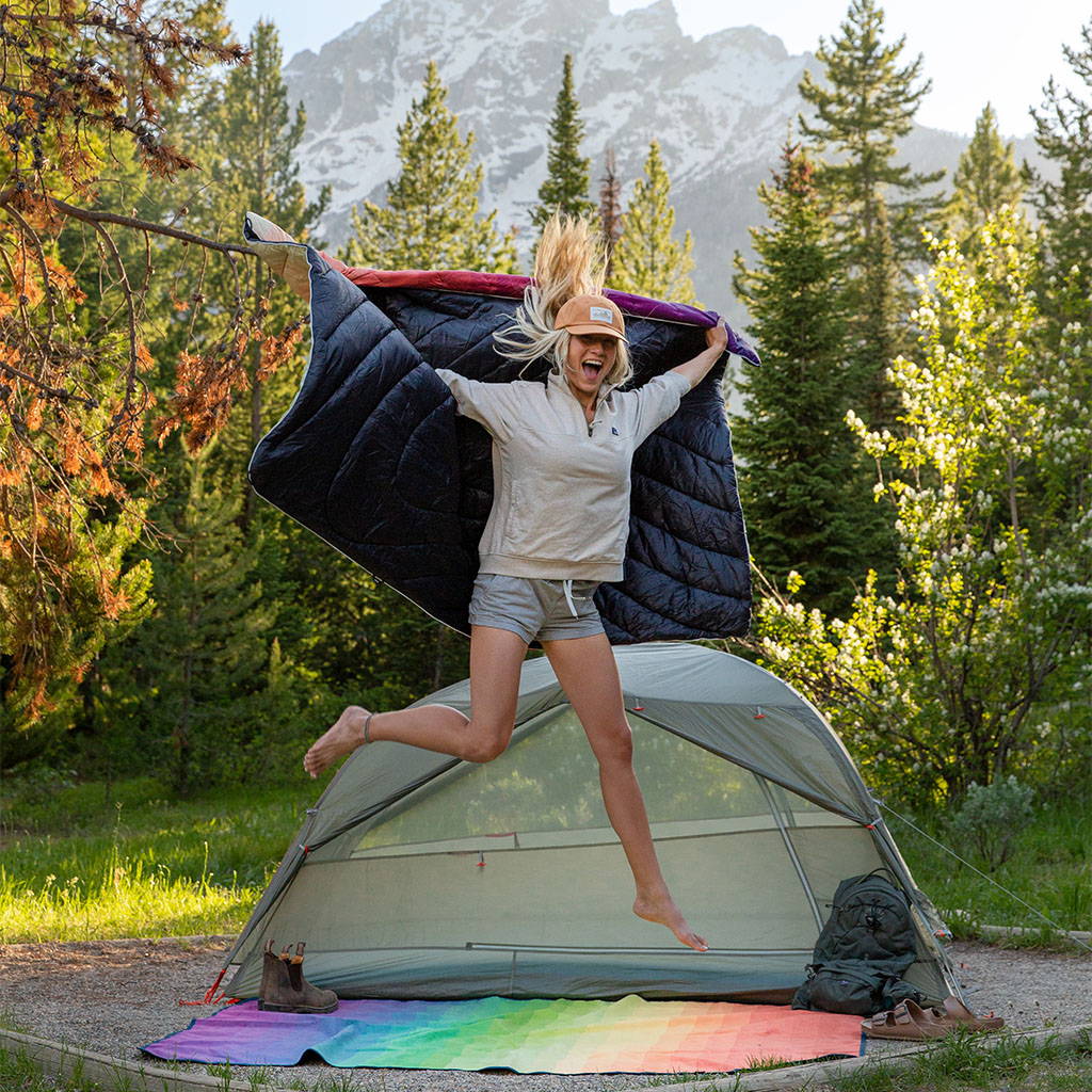 Excited girl with camping gear. She jumps up with her Rumpl blanket in hand, her tent and Everywhere Mat visible behind he