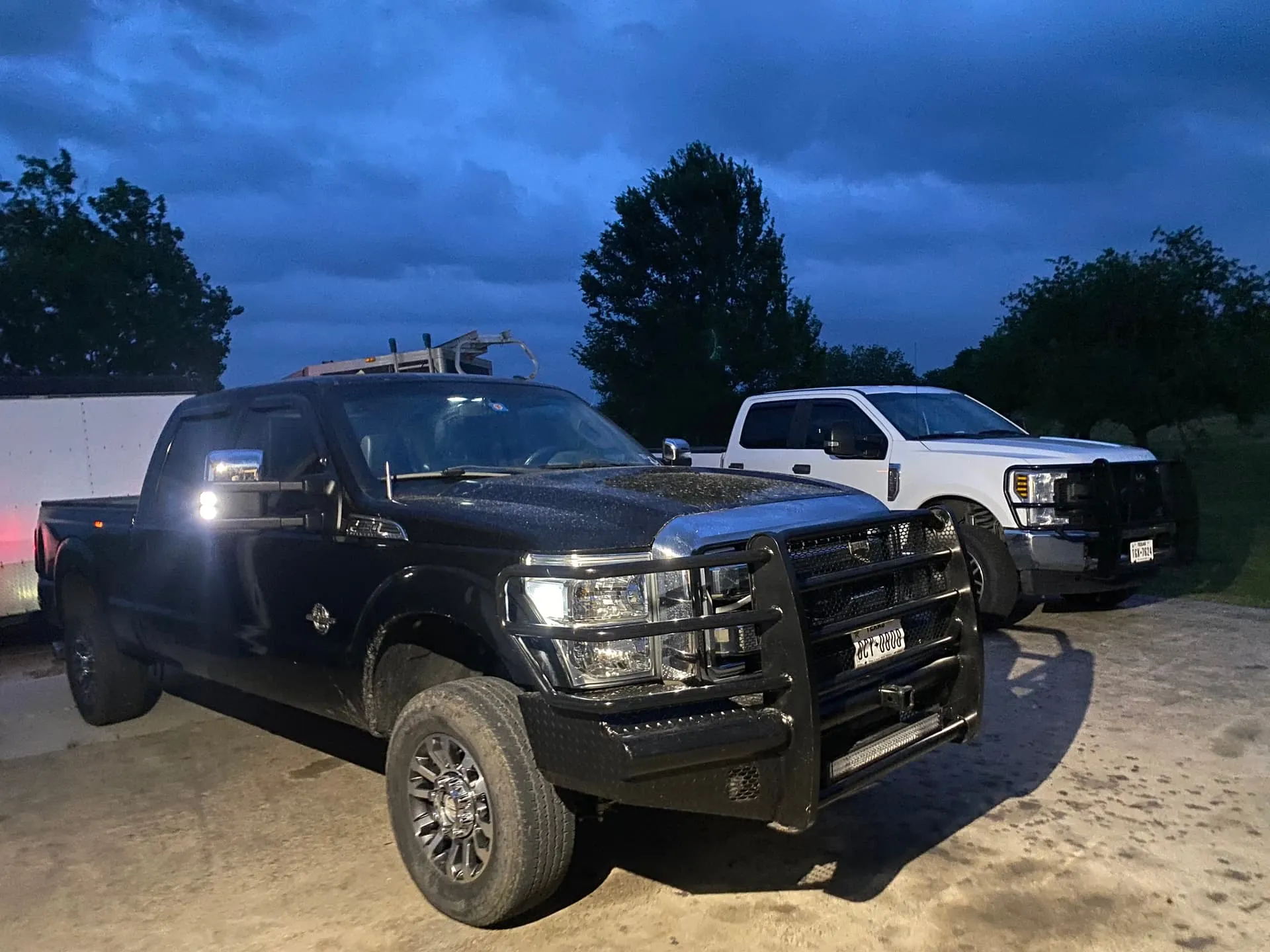 Two Ford Super Duty pickup trucks, black and white, parked at twilight with heavy-duty front brush guards