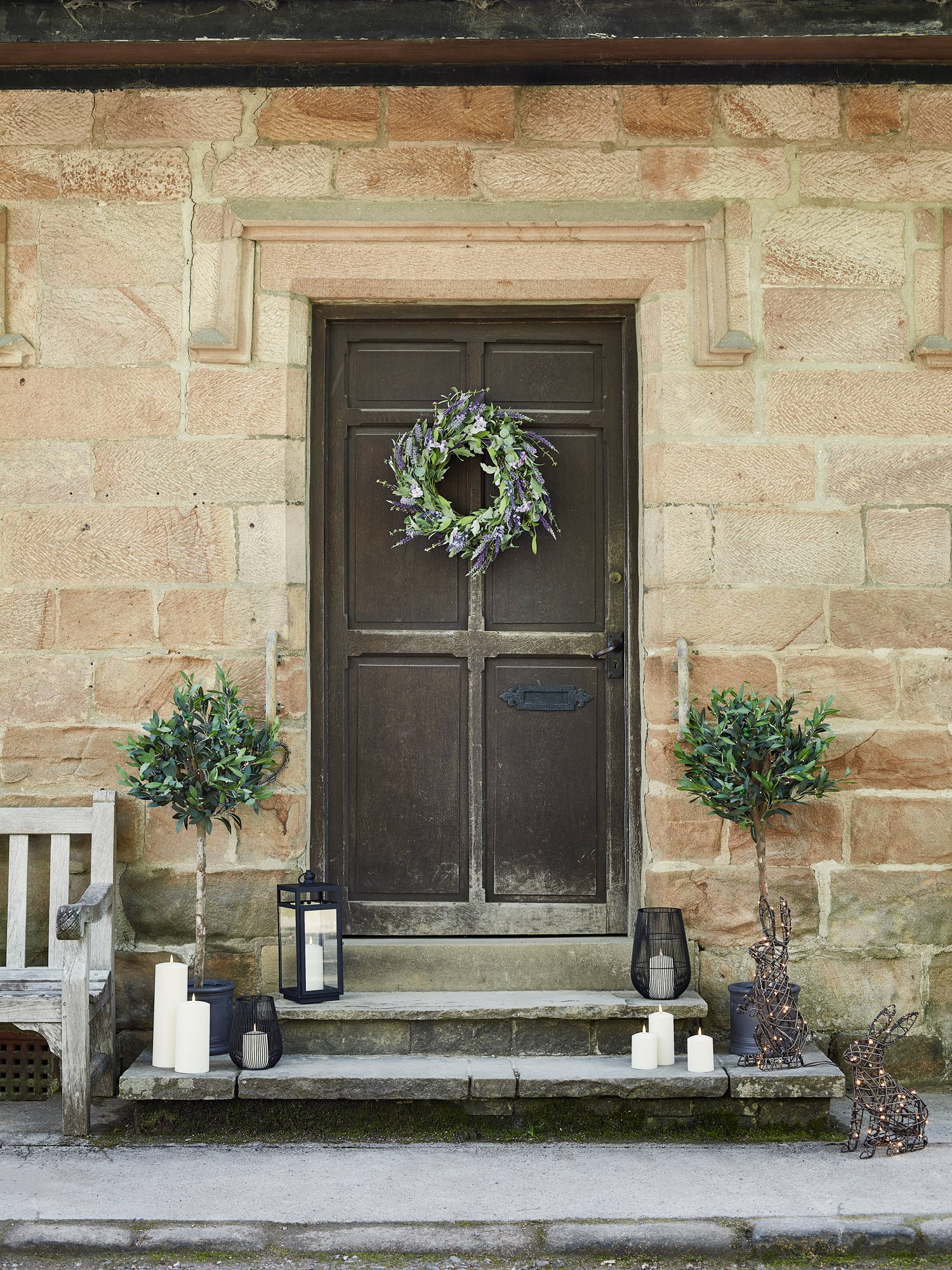 Doorstep with spring lavender wreath on door with assortment of black lanterns and outdoor candles sat on steps with rattan bunnies