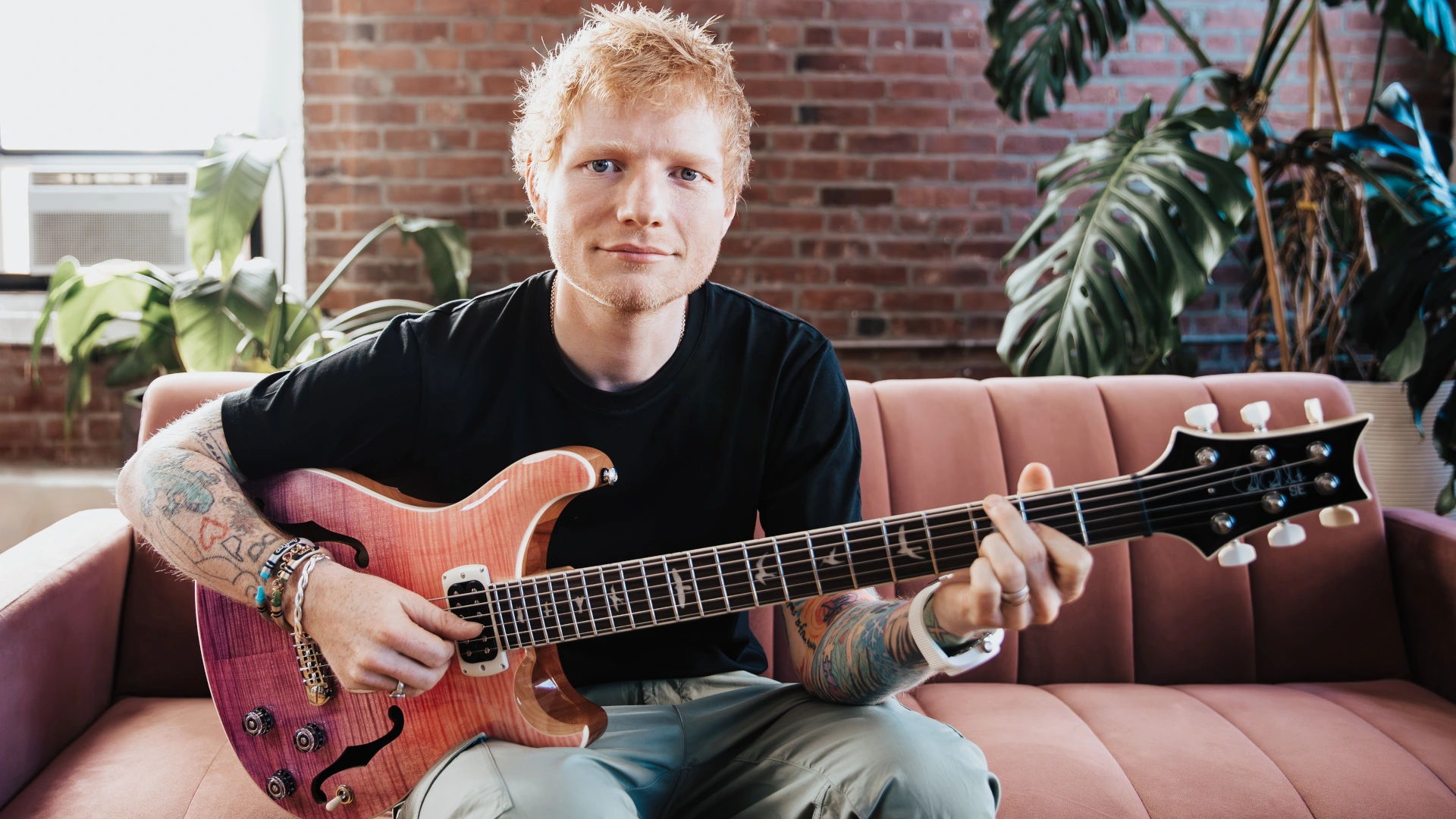 Ed Sheeran sitting on a couch playing an electric guitar in a cozy setting.