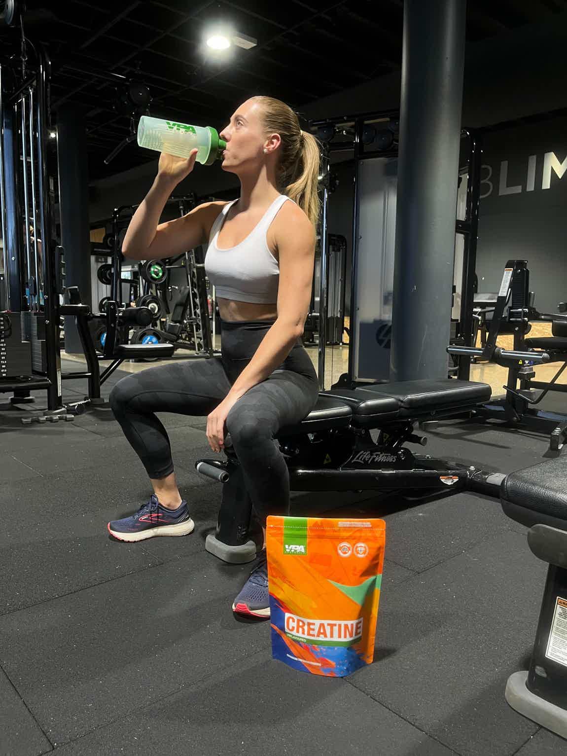 Woman drinking from a bottle in a gym, with a supplement container nearby.
