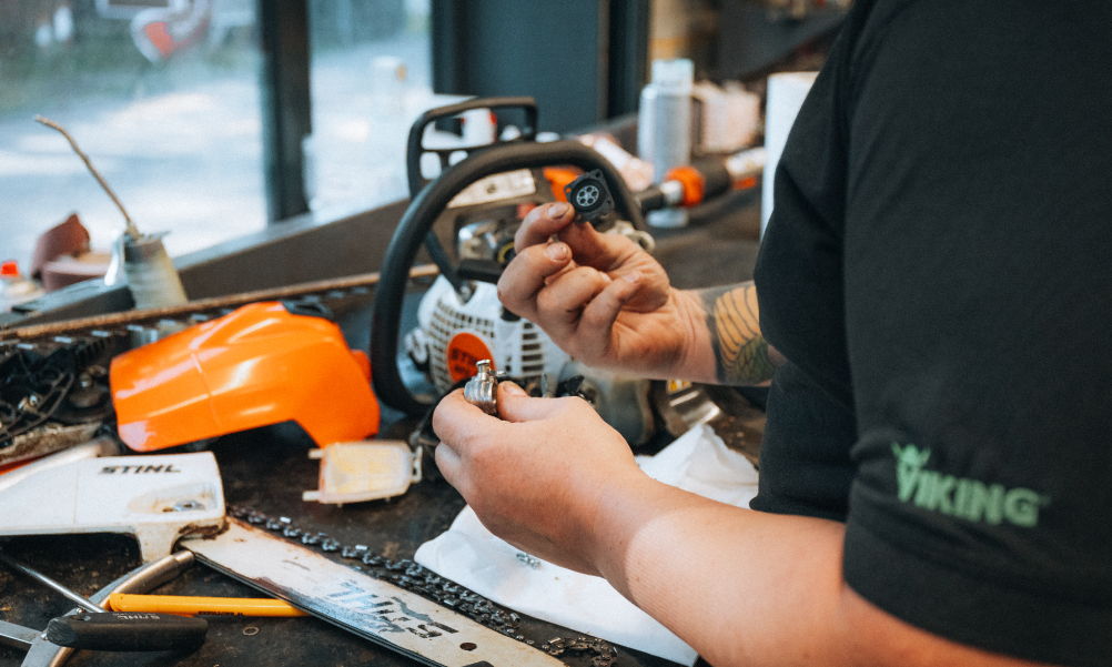 Person repairing a chainsaw, holding tools and parts on a workbench.