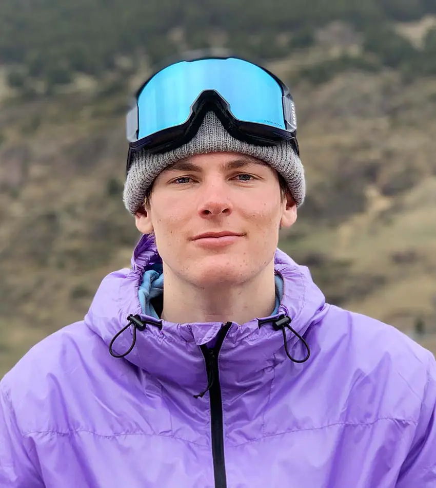 A young man wearing a purple jacket and blue ski goggles outdoors.