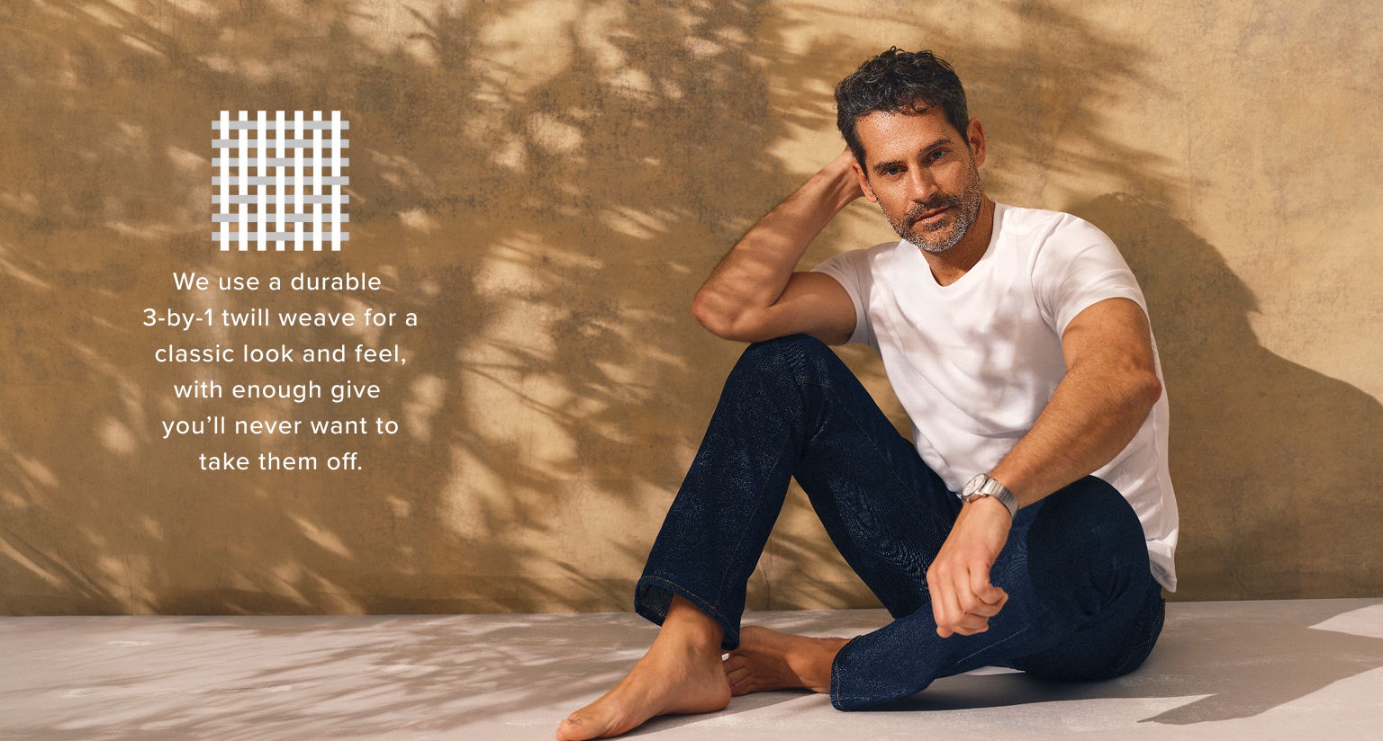 Man sitting casually against a wall, wearing a white shirt and dark pants.