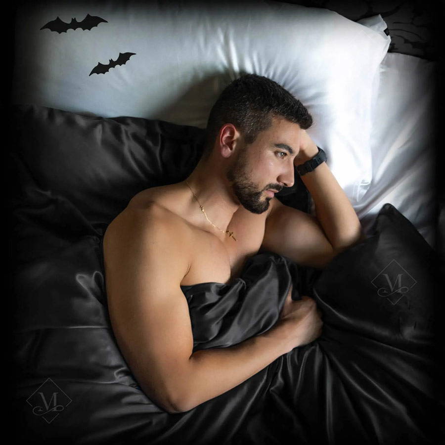 man in bed with dark silk sheets and halloween bats on the pillowcase