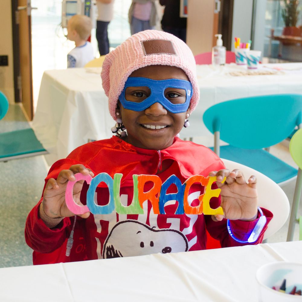 Small Child wearing a superhero mask holding up a colorful sign that says Courage