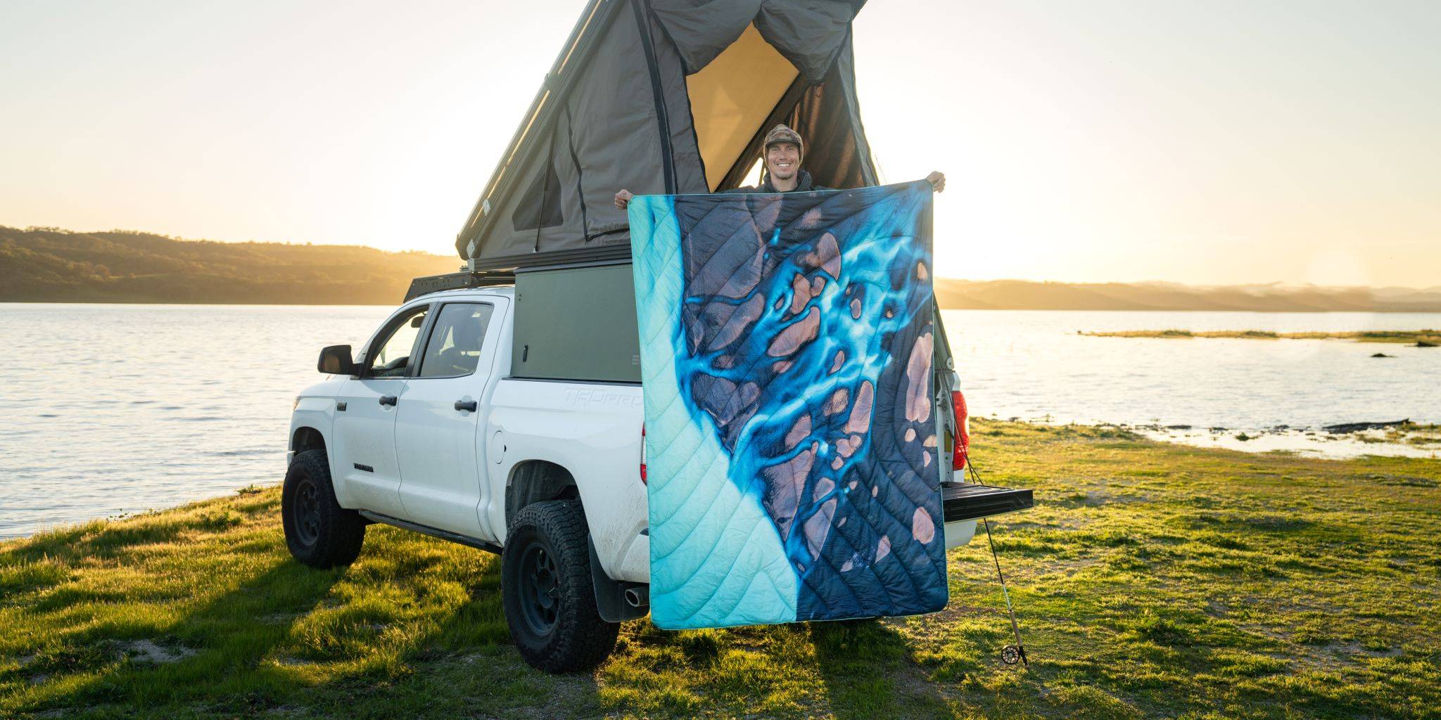 Chris Burkard holding up the Rumpl Original Puffy Blanket - Thjorsa while standing by a white pickup truck with an open rooftop tent near a calm lakeshore with grassy terrain during sunset.