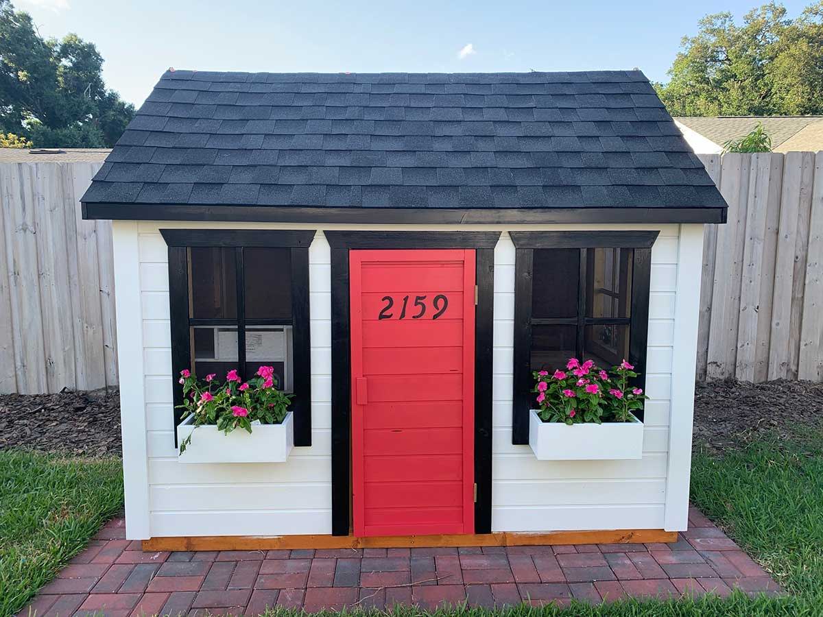 Wooden playhouse with a black roof and white flower boxes with pink flowers by WholeWoodPlayhouses