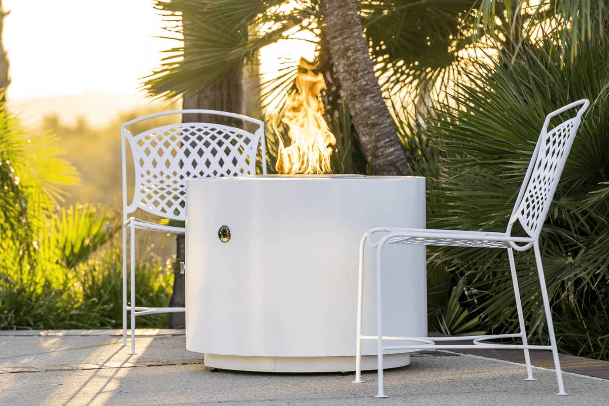 Boxhill's Beverly Powder Coated Fire Pit in white is burning bright in a tropical patio surrounded by two geometric metal white chairs. 