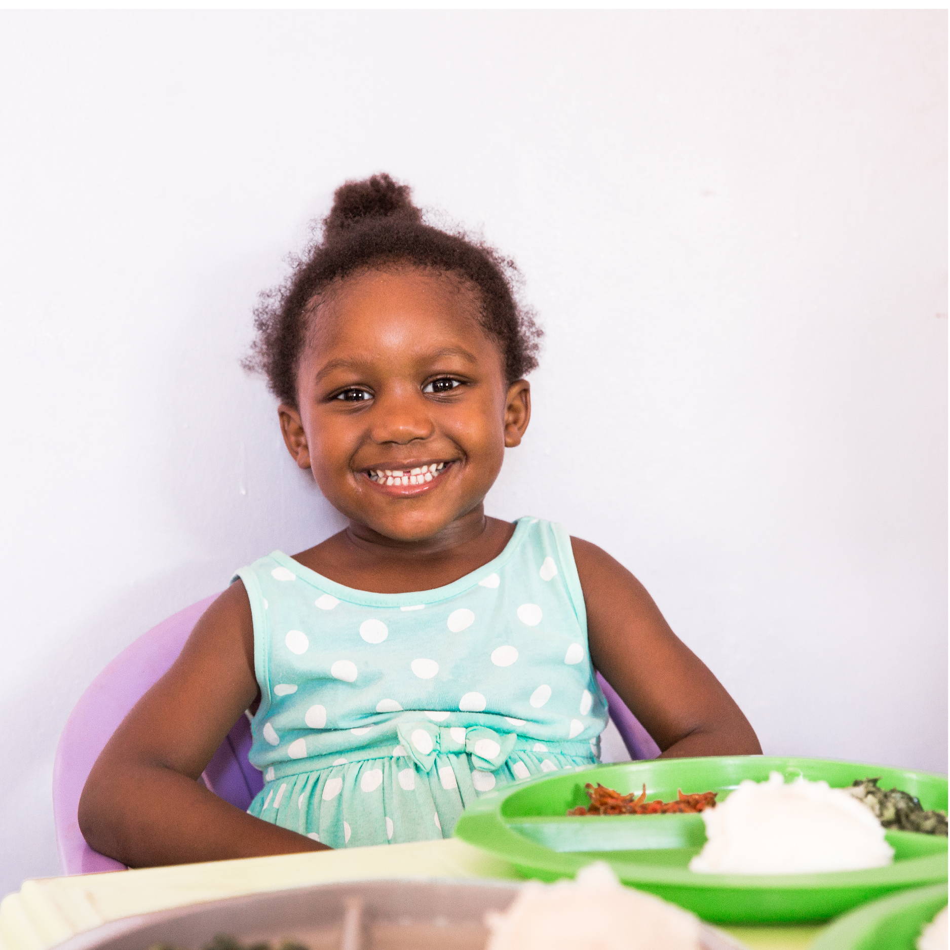 A small Zambian girl in a blue polka dotted dress gives a big smile, showing her teeth, as she sits to eat lunch. 