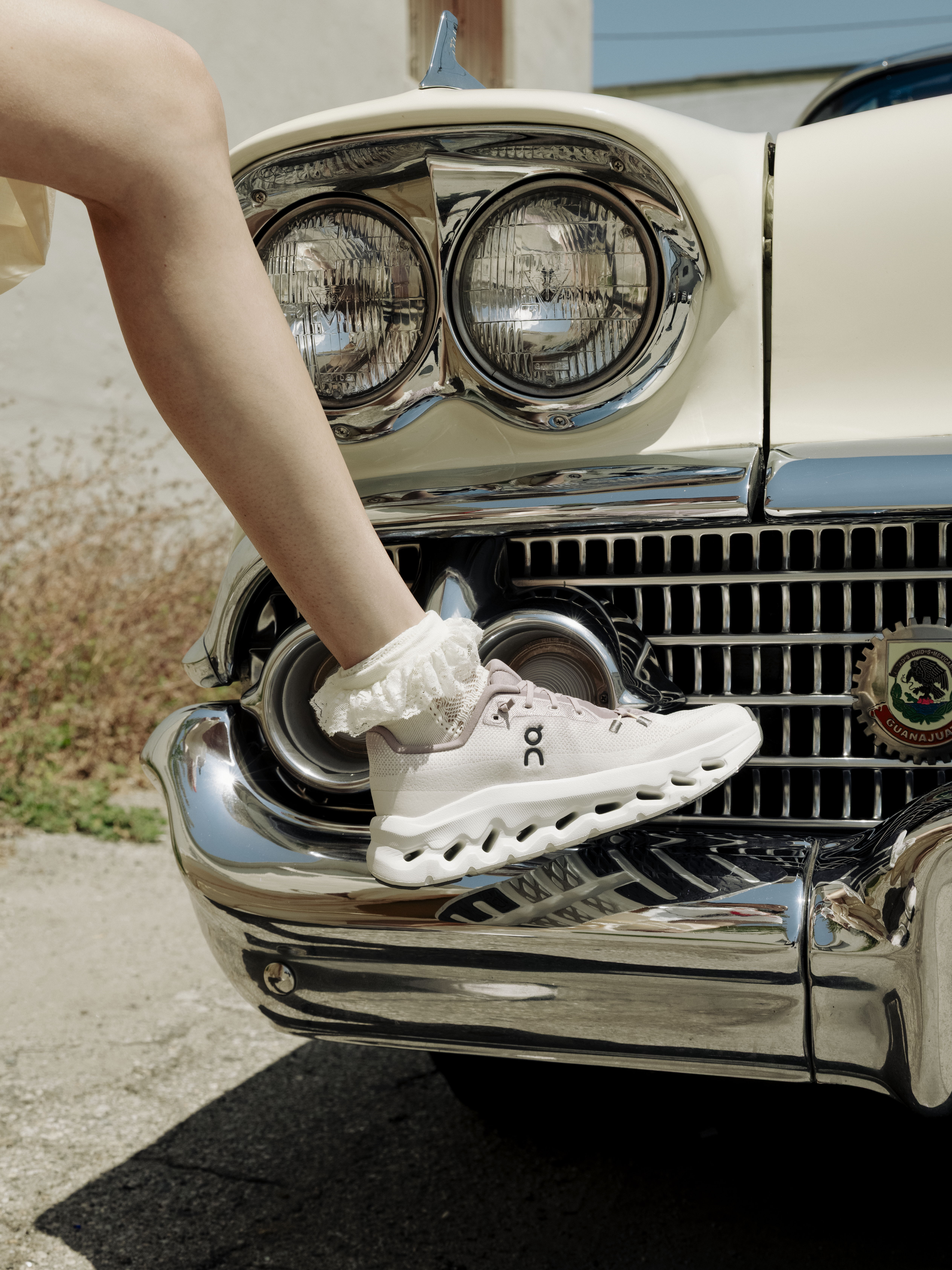 Person's foot in sneaker resting on a vintage car's chrome bumper.